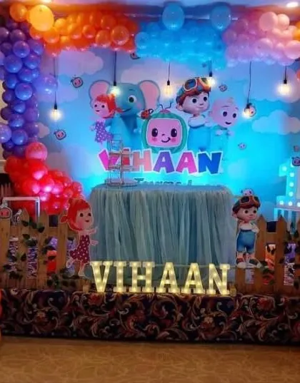 A Cocomelon party setup featuring a sky-blue backdrop with clouds and hanging light bulbs. The decoration includes multiple character props and colorful balloon clusters.