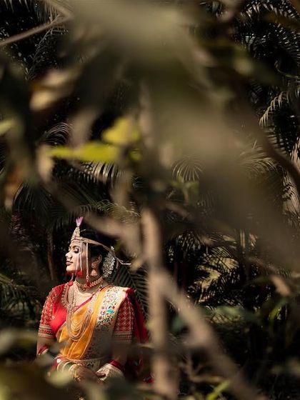 A bride is captured through the foliage, creating a natural frame. The sunlight catches her traditional Maharashtrian attire, giving her a serene and almost ethereal glow.