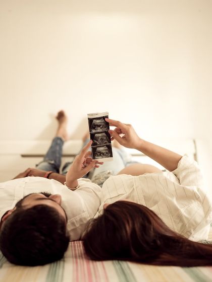 A creative and relaxed indoor shot. The couple lies on the bed, looking up at the sonogram pictures they hold, sharing a private moment of wonder.