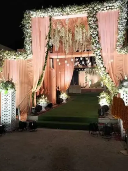A breathtaking nighttime wedding entrance that exudes luxury. The design features soft peach draping, a canopy of white flowers, hanging chandeliers, and spotlights to create a warm and inviting pathway for guests.