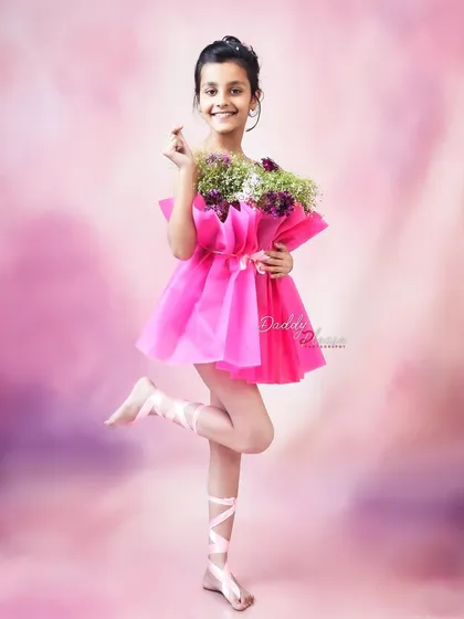 A smiling girl poses like a ballerina in her creative pink floral dress. The soft pink background and her joyful expression make this a delightful portrait.