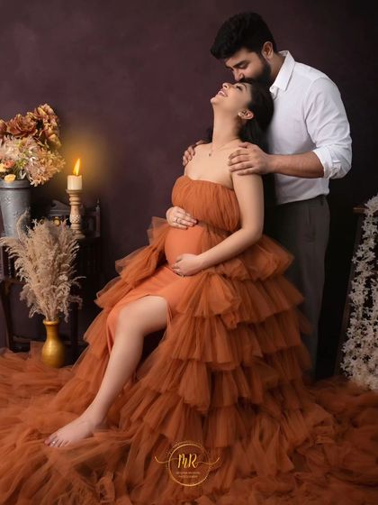 A joyful and romantic pose. The partner kisses his wife's forehead as she leans back happily, seated in a beautiful orange gown in a candlelit setting.