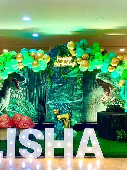 For older kids, I can create more thrilling themes like this dinosaur party. The backdrop features realistic T-Rex graphics, surrounded by a jungle of green and gold balloons and personalized marquee letters for Alisha.