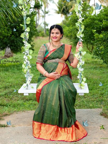A beautiful outdoor portrait of a mother-to-be in a green and red silk saree, seated on our floral swing.