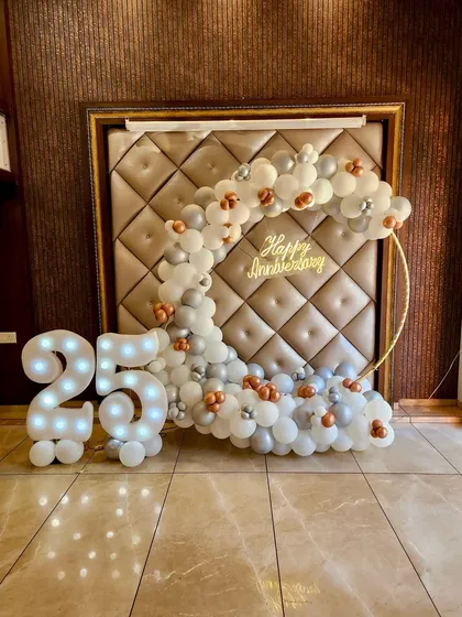 Celebrating a milestone anniversary? This elegant setup for a 25th anniversary features a crescent-shaped balloon arrangement in white, silver, and rose gold on a golden ring. The lit-up marquee number "25" and a "Happy Anniversary" neon sign make it a perfect photo spot.