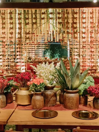 A reception dinner setting that combines rustic wooden tables with elegant brass plates and vibrant red and green floral arrangements, creating a warm and inviting atmosphere.