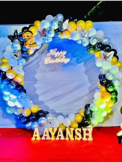 A colorful circular balloon arch for Aayansh's 2nd birthday. This setup uses a mix of blue, yellow, white, and black balloons, with butterfly accents and the name displayed in wooden letters.