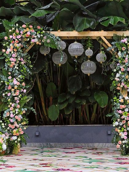 A beautiful floral arch against a backdrop of giant green leaves. The pastel-colored flowers and hanging lanterns create a soft and romantic photo spot.