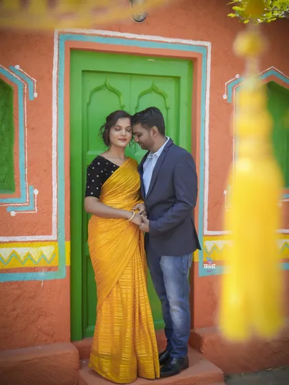 An intimate moment between the couple, framed by the doorway of a colorful, rustic village house set. The foreground blur adds a dreamy quality.