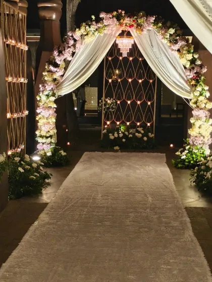We designed this elegant entrance for a Roka ceremony. The walkway features a white carpet, a floral arch with pastel flowers and drapes, and candle-lit stands, creating a romantic and welcoming path.