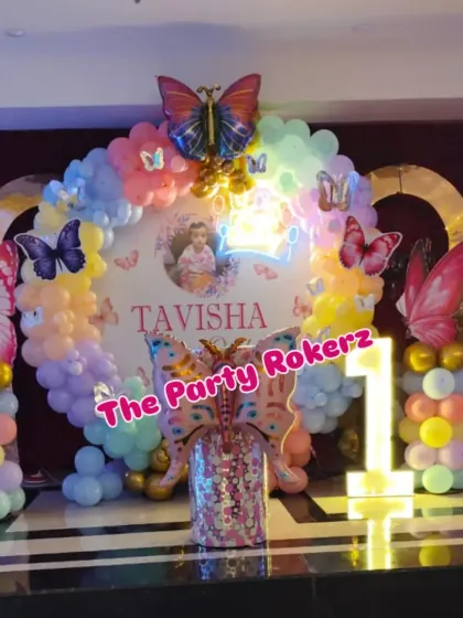 A nighttime view of the butterfly birthday setup, showing how the neon sign and marquee number light up the space. The pastel balloons and butterfly details look magical under the lights.