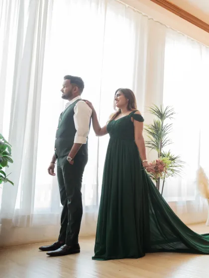 A beautifully composed shot by a large window. The bride, in her elegant green gown, places a hand on the groom's shoulder as they both look out, creating a reflective and serene mood.