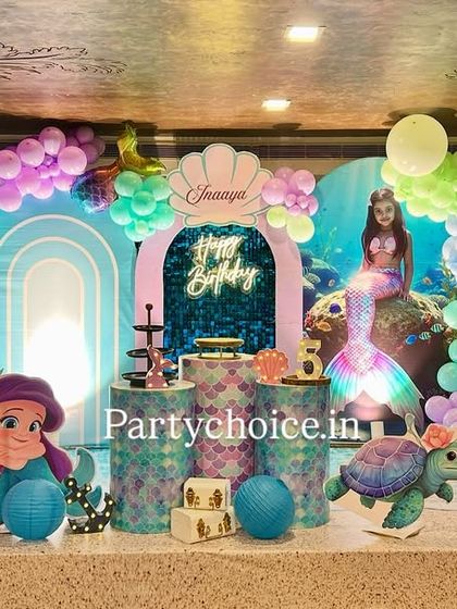 An enchanting mermaid-themed birthday party. The underwater scene is created with a blue and purple balloon arch, a shimmering backdrop, and cutouts of Ariel, a sea turtle, and coral.