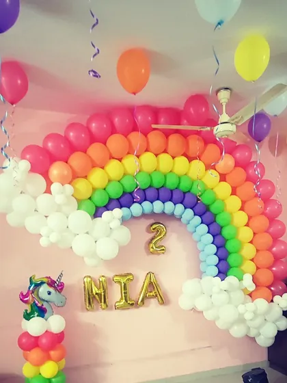 A beautiful rainbow balloon arch against a pink wall for a second birthday. This decoration includes balloon clouds, a unicorn balloon column, and the name "MIA" in gold foil letters below the rainbow.