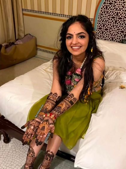 A smiling bride sitting on a bed, proudly showing her complete bridal mehndi on hands and feet.