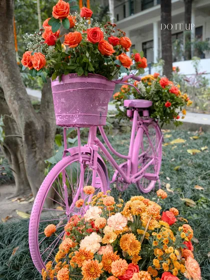 A whimsical decor element for the Haldi by the Ganges. A vintage-style bicycle is painted pink and filled with baskets of bright orange flowers, adding a playful and photogenic touch to the outdoor setting.