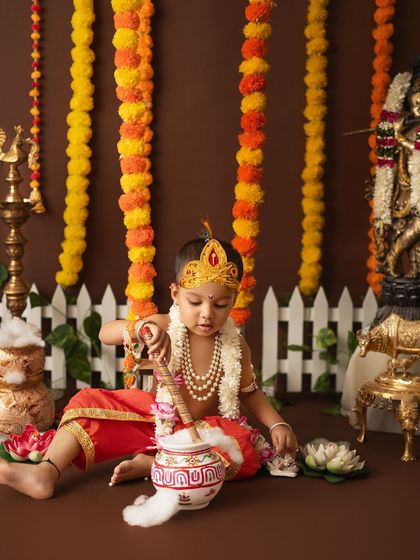 A playful shot of a little boy as Krishna, churning a pot of "butter" in a setup complete with a white picket fence and marigolds.