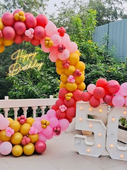 A vibrant and cheerful 50th birthday celebration. This outdoor setup features a massive circular balloon arch in shades of pink, red, and yellow, a "Happy Birthday" neon sign, and large "50" marquee numbers, creating a perfect, joyful photo opportunity.