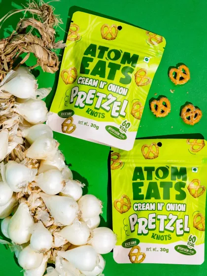 A vibrant shot of Atom Eats Cream 'n' Onion pretzels with a braid of fresh pearl onions against a bright green background. This composition creatively links the product to its core ingredient.