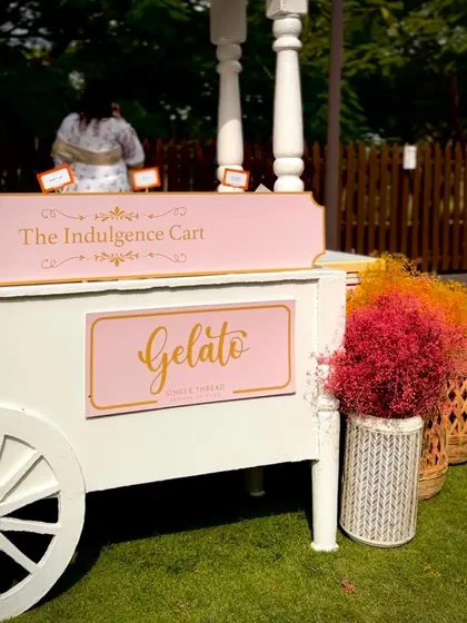 Our beautiful white and pink gelato cart, branded "The Indulgence Cart," is set up on a lush green lawn. This image showcases the charming and professional setup we bring to events.