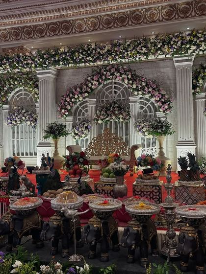 A majestic stage design inspired by classical architecture, with large white pillars, arches, and green floral walls. The offering table features a long line of black elephant props.