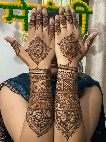 A beautiful pose showcasing a symmetrical bridal design. The central mandalas on the back of the hands are the focal point, complemented by heavy, traditional work on the forearms.