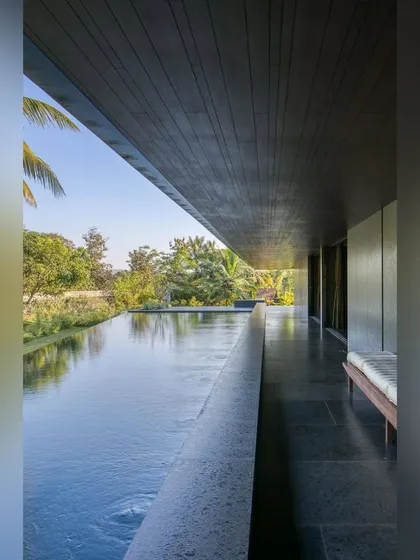 A long, linear infinity pool runs alongside the veranda of this Alibaug home. The dark stone and reflective surface mirror the sky and surrounding trees, making the pool an integral part of the landscape.
