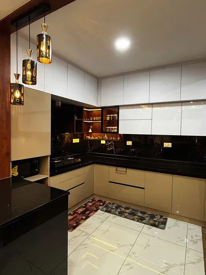 This acrylic kitchen combines glossy white and beige cabinets with a black granite countertop for a timeless look. The hanging pendant lights over the breakfast counter add a touch of warmth and style.