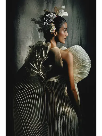 A profile shot showcasing the back of a pleated, sculptural gown by Vaishali S. The side lighting beautifully illuminates the garment's form and the intricate headpiece.