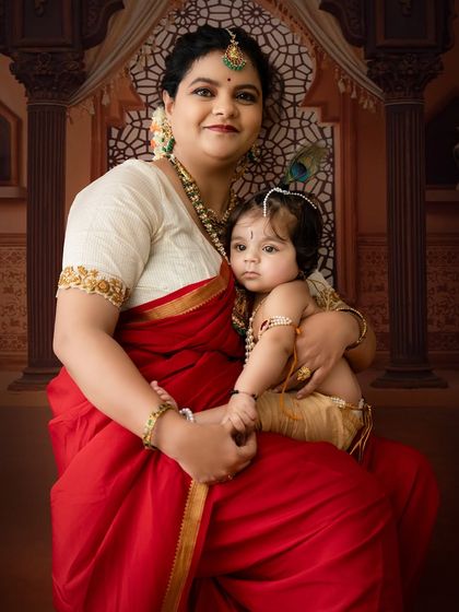 A classic seated portrait of a mother holding her little Krishna, showcasing the intricate details of their traditional outfits and the temple-themed set.
