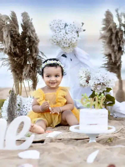 A perfect first birthday portrait on the beach. This shot captures the full boho-themed setup with the cake, "One" sign, and the ocean in the background.