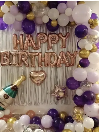 A birthday decoration with a silver fringe backdrop. The setup includes a balloon garland in purple, gold, and white, and a "Happy Birthday" foil banner.