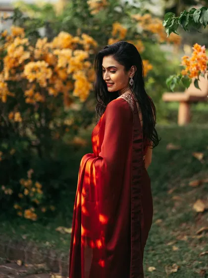 A beautiful portrait of a woman in a red saree, smiling as she stands in a garden, with soft sunlight highlighting her hair.