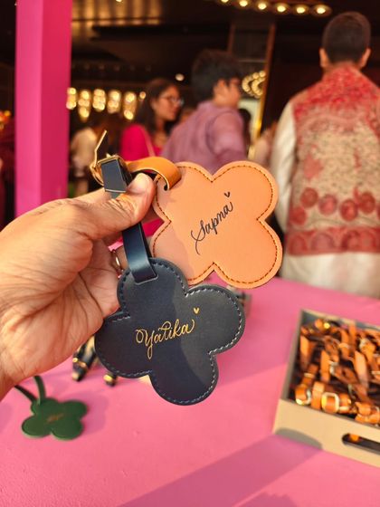 Holding up two personalized luggage tags at a Mehendi event. Each name is written by hand, offering guests a truly unique and personal party favor.