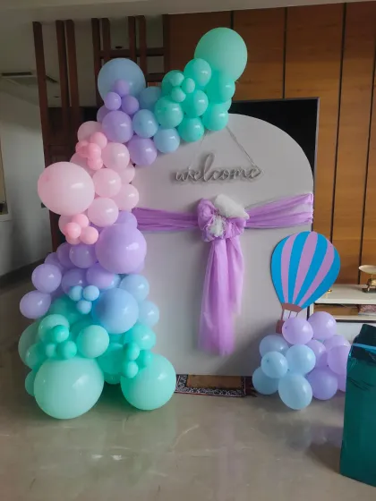 A modern and elegant naming ceremony decoration with a pastel balloon garland on a white circular backdrop. A small hot air balloon prop adds a whimsical touch.