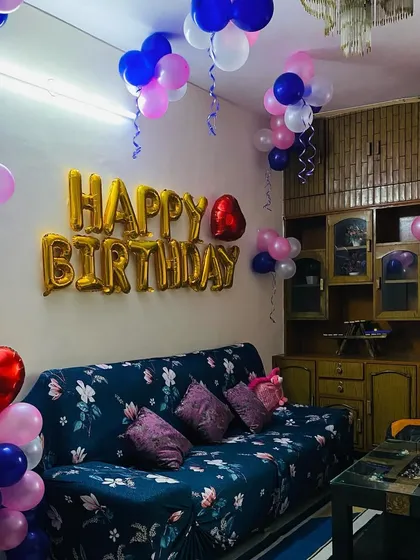 A cozy living room decorated for a birthday with pink, blue, and white balloons on the ceiling and a gold "Happy Birthday" foil banner.