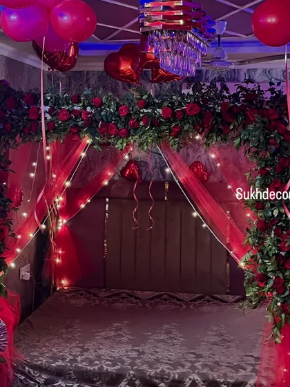 A detailed shot of the floral canopy over the bed. You can see the arrangement of red roses, heart-shaped foil balloons, and the soft glow of the fairy lights against the red fabric.