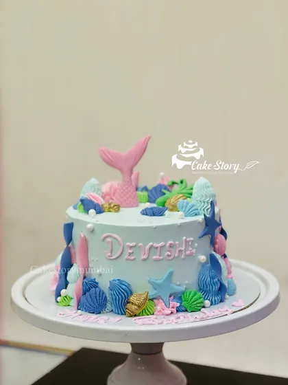 A lovely mermaid-themed cake. The light blue cake is decorated with a pink mermaid tail and a variety of colorful, edible seashells and starfish.