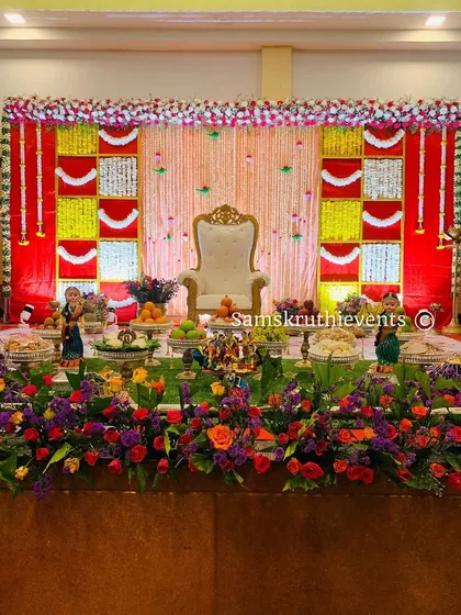 A classic Seemantha decor setup with a red, yellow, and white color scheme. The stage includes a central throne, traditional dolls, and a flowerbed, making it a perfect setting for the ceremony.
