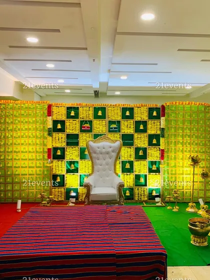 A wide-angle view of a traditional baby shower stage. The backdrop features a checkerboard of betel leaves and marigolds, with a grand white throne as the centerpiece.