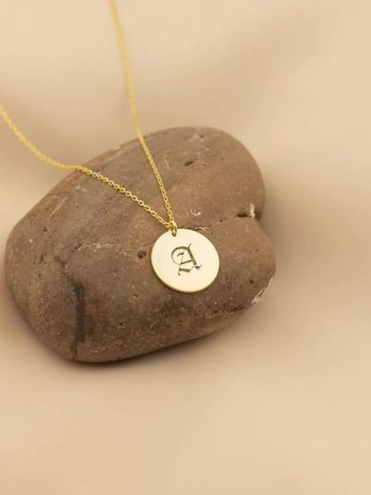 A gold round pendant engraved with a gothic-style initial 'A'. This product shot shows the classic elegance of an initial pendant, perfect for gifting or self-purchase.