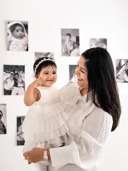 A beautiful mother-daughter moment. The background wall of photos adds a personal touch, celebrating the family's journey leading up to this first birthday shoot.