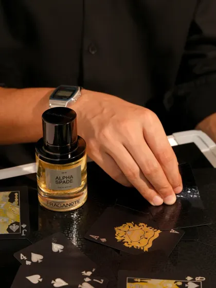 A lifestyle shot for a men's fragrance, featuring a hand model dealing cards. This places the product in an aspirational context of sophistication and gamesmanship.