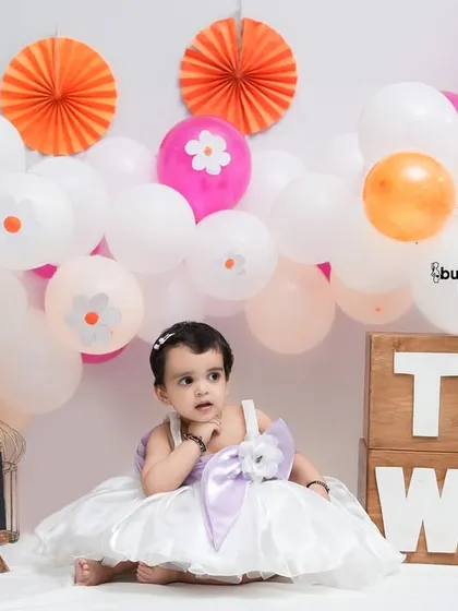 A thoughtful pause during her second birthday photoshoot. The colorful setup with balloons and paper fans creates a cheerful and celebratory atmosphere.
