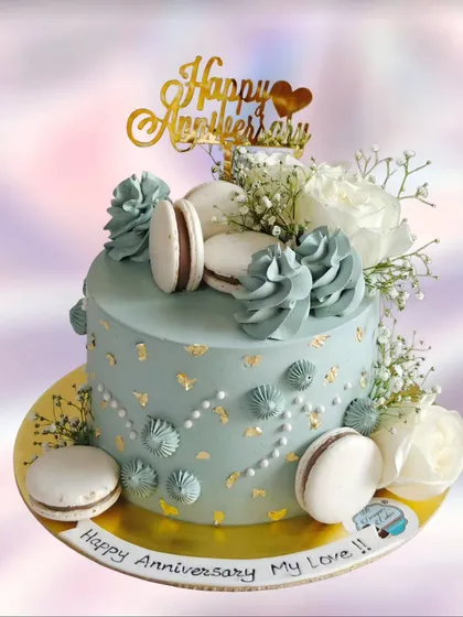 An elegant anniversary cake in a dusty blue shade, adorned with macarons, fresh white roses, and gold leaf. A sophisticated choice for a romantic celebration.
