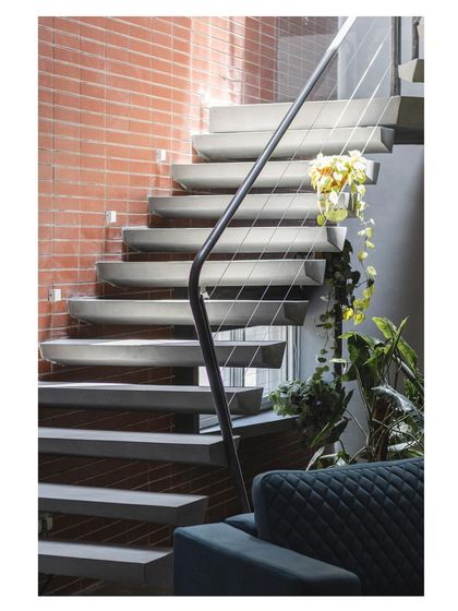 The full context of the floating staircase within a home, set against a warm brick wall. The thin steel cables provide safety without visually obstructing the sculptural form.