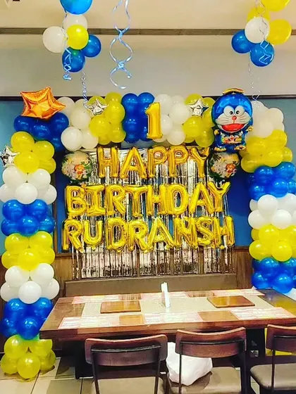 A Doraemon-themed first birthday setup with a balloon frame, silver fringe curtain, and the name 'RUDRANSH' in gold foil letters.