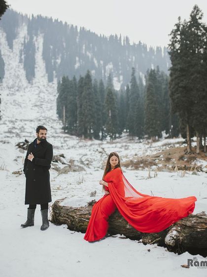 A stunning composition from a Kashmir pre-wedding shoot. The bride's red gown drapes over a fallen log in the snow, creating a powerful contrast with the majestic, snow-covered forest behind them.