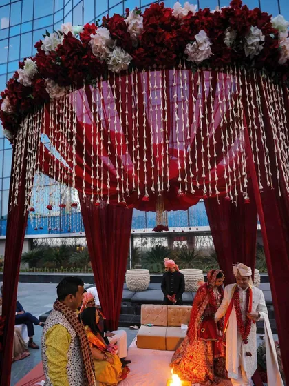 A classic round mandap design for a traditional Hindu wedding. The structure is draped in rich red fabric, with a canopy of red and white flowers and hanging tuberose strings.
