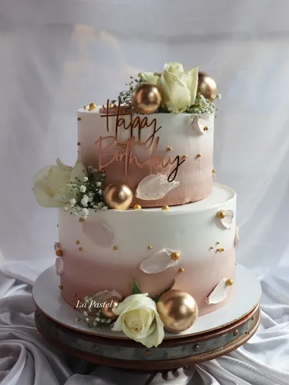 A beautiful two-tier cake with a soft pink ombre effect. Gold spheres, white roses, and delicate petal details make this a perfect choice for a sophisticated birthday or engagement.